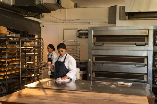 Female bakers cleaning prep table in chef coats using sprayer and organizing rack trays in bakery