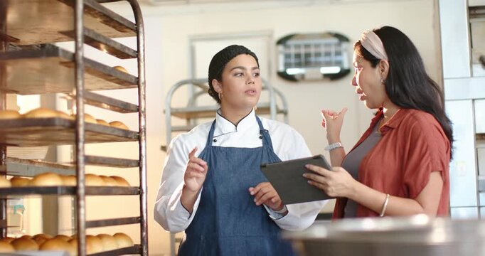 Diverse women manager tapping tablet cueing chef in coat pointing at rack checking trays at bakery