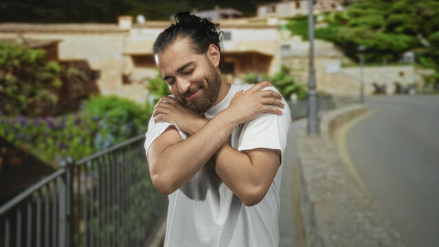 Young arab hispanic man hugging his arms across chest on a street near railing and buildings; self care serenity.