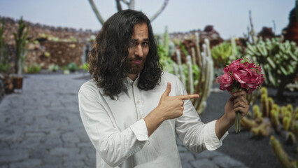 Fototapeta premium Man smiles and points finger to bouquet of pink flowers in park among cacti and stone path; romance playful charm.