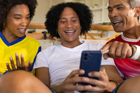 African friends sitting on couch in living room, laughing while sharing smartphone and jerseys