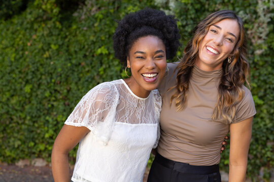 Smiling diverse female friends standing by ivy wall, wearing white lace and beige turtleneck