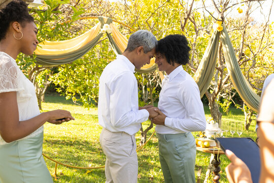 African American men standing holding hands touching foreheads in garden with drapery, cake