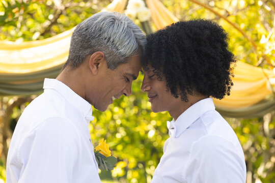 Male pair touching heads under gold arch in park, in white shirts, yellow boutonniere, white bowtie