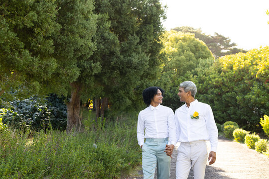 Two African American men holding hands walking sunny gravel path with yellow flower pin, copy space
