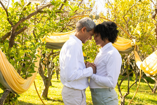 African American men touching foreheads holding hands in garden under yellow arch in white shirts