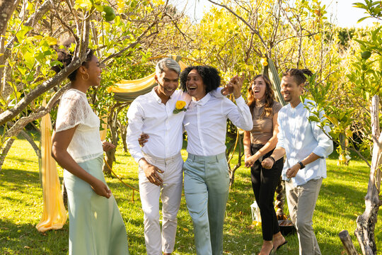 Two African American men walking arm-in-arm in orchard under yellow fabric arch wearing boutonniere