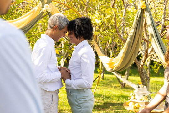 African American men pressing foreheads, clasping hands at ceremony in garden, cake tray nearby
