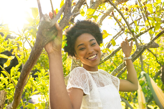 African American woman holding branches smiling in sunny orchard wearing white lace dress watch