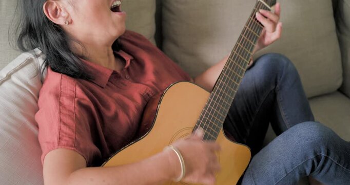 Woman is settling on light couch, starting to strum acoustic guitar and singing while relaxing