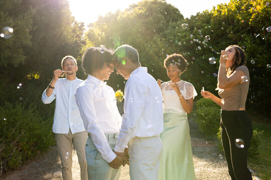 Two African American men holding hands touching foreheads vowing as guests blowing bubbles on path