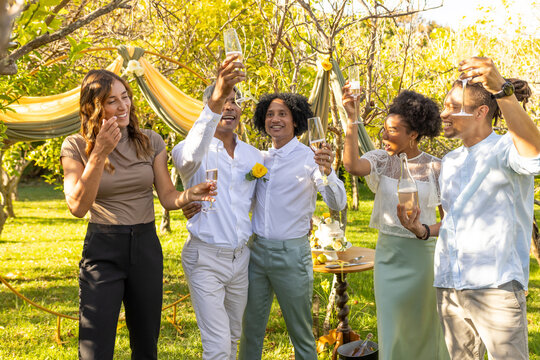 Diverse friends toasting after cake, couple linking arms in white shirts, yellow pins at garden