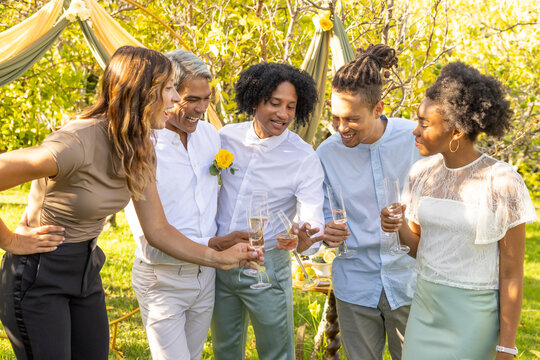 Diverse friends clinking flutes under draped arch in garden, wearing yellow boutonniere