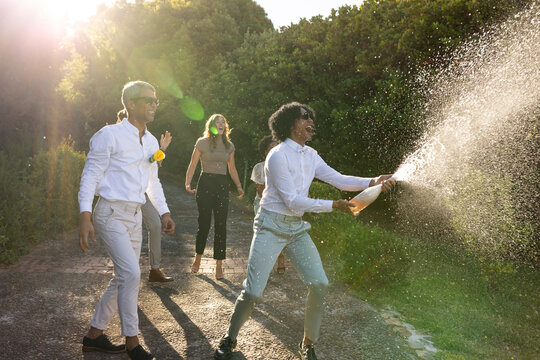 Diverse friends walking paved path celebrating, man spraying champagne, others laughing
