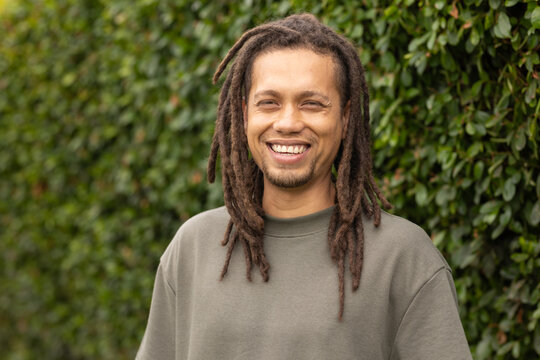 African American man standing smiling by green hedge wearing olive crewneck with dreadlocks