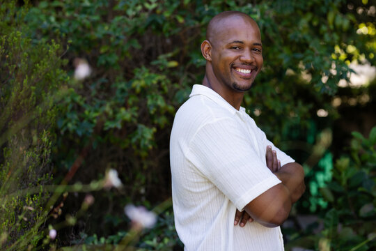 Adult African American man posing arms crossed in light collared shirt near yard shrubs, copy space