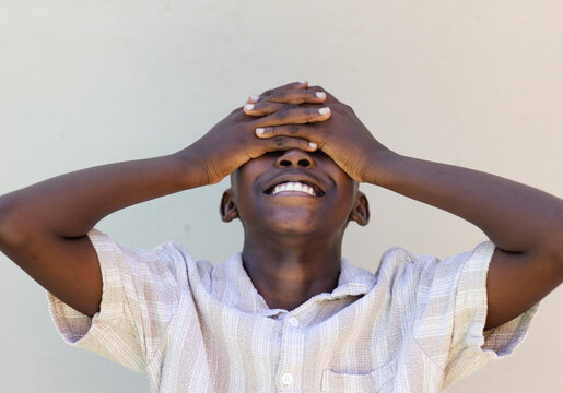 African male youth covering eyes with hands and smiling in short sleeve shirt against beige wall