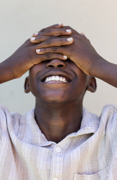African youth male tilting back covering eyes smiling wearing light collared shirt by beige wall