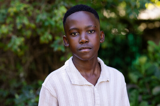African adolescent male standing and facing camera in garden, wearing light beige collared shirt