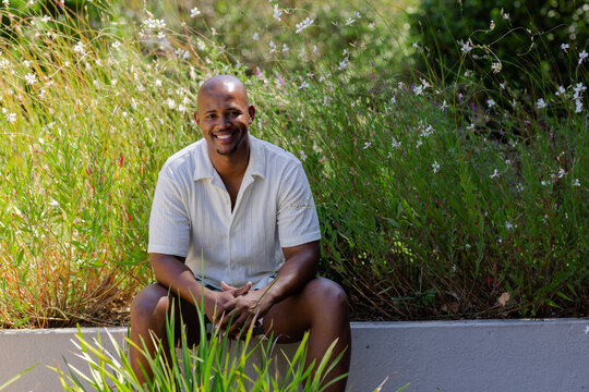 African man sitting on concrete planter wall in garden with ornamental grasses and flowers