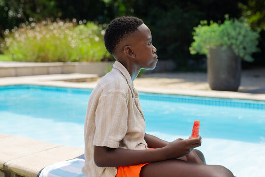 African American male youth sitting on pool coping holding red popsicle at backyard pool on towel