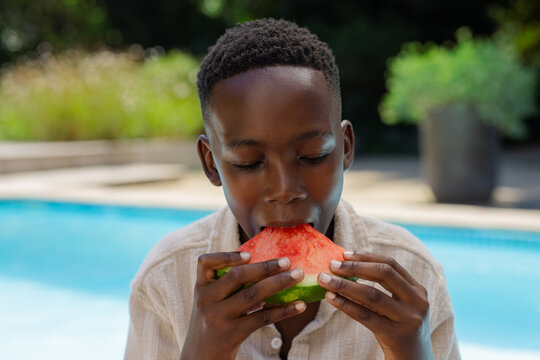 African adolescent male sitting at backyard pool wearing beige shirt, biting large watermelon slice