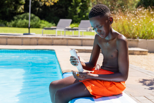 African teenager sitting poolside in orange trunks using white sunscreen on blue towel, copy space