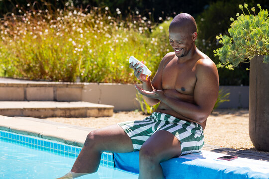 African American man sitting poolside applying sunscreen from bottle in trunks, phone, copy space
