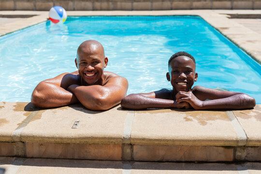 African father and son leaning on stone coping at pool, shirtless and smiling by beach ball