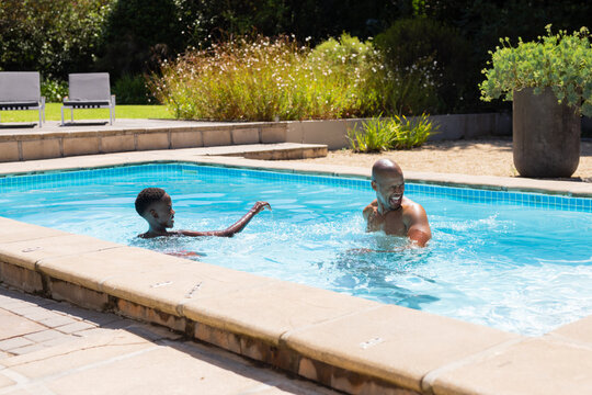 African father and adolescent son laughing and splashing in backyard pool with stone coping