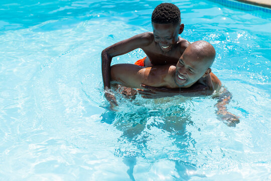 African adult and youth males in trunks piggybacking in blue pool, splashing sunlit water ripples