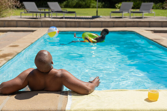 Sunny day causing shirtless man leaning poolside watching teen floating on green ring, drink