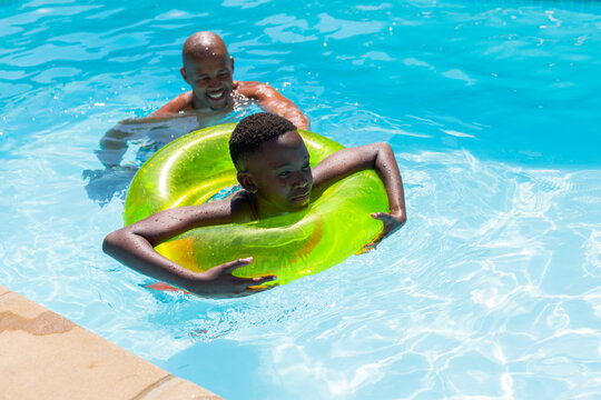 African American man supporting boy in neon green ring, floating near tan pool deck, wearing trunks