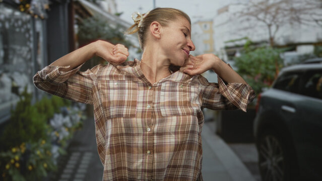 Woman stretching arms wide on a city street with closed eyes and slight smile while wearing plaid shirt and reaching outward; tired recovery.