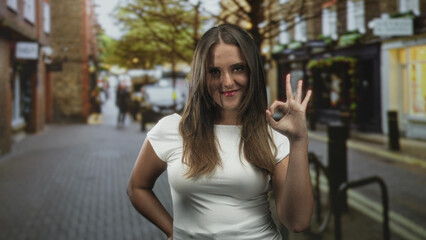 Fototapeta premium Young woman making ok hand gesture with hand on hip wearing a white top on a street lined with storefronts and trees; confidence approval.