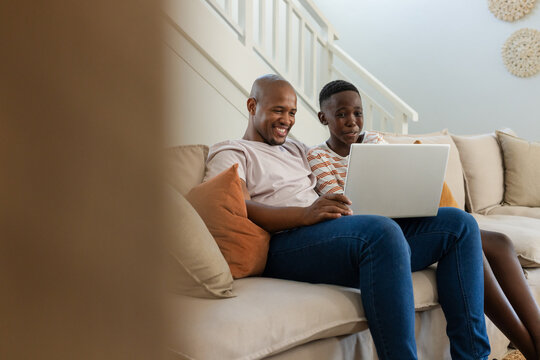 African father and teen son sitting on beige sofa at home using silver laptop together, smiling