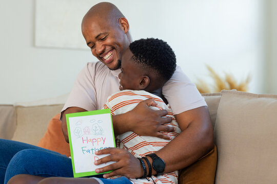 African American father and child hugging on sofa at home, holding green bordered Father's Day card