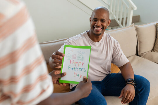 Vertical video: African American dad smiling, receiving Fathers Day card from child on sofa