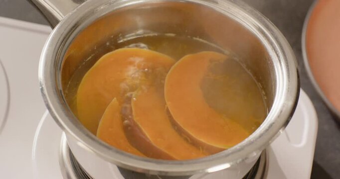 As white burner is heating, steel saucepan is simmering three orange squash slices softening edges