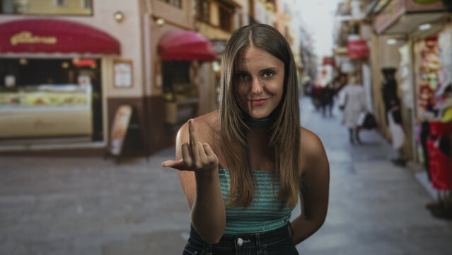 Woman shows middle finger toward camera on a narrow pedestrian street with shop awnings and blurred passersby; defiance.