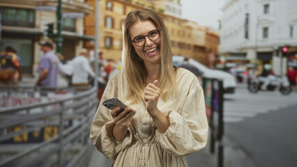 Obraz premium Blonde woman smiling and tapping smartphone with both hands, wearing glasses and beige dress on a busy city street crosswalk; relaxed joy.