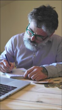 Worried mature man with a beard calculating his expenses, writing on a notepad and feeling stressed about his financial situation