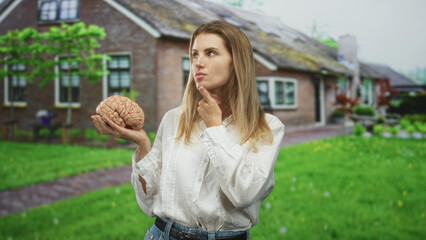 Fototapeta premium Blonde woman holding brain prop, finger on chin while posing in a cottage front yard with green lawn and path; curiosity reflection.