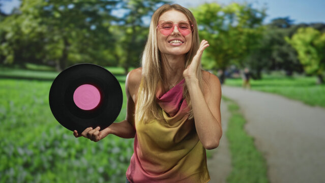 Blonde woman holding a vinyl record, hand to cheek singing on a forest path with pink sunglasses and colorful top; nostalgia joy celebration.