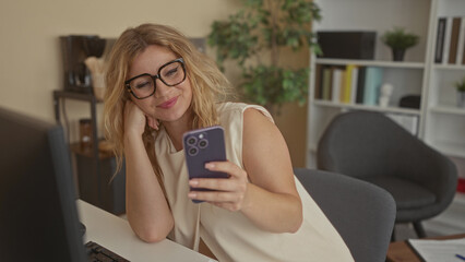 Fototapeta premium Woman wearing glasses holding smartphone, smiling while seated at a desk in an office interior beside a computer monitor; happiness work balance.