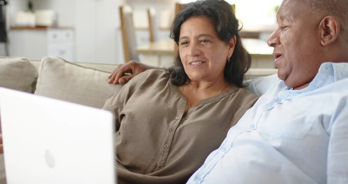 Senior couple sitting on beige sofa in living room, watching white laptop, conversing about content