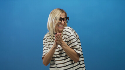 Fototapeta premium Woman middle aged blonde clapping hands and smiling in studio against blue backdrop wearing striped t shirt and glasses; joy celebration.