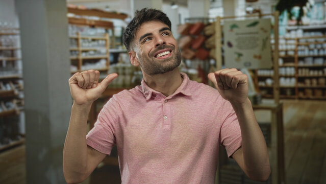 Man wearing pink shirt smiling and pointing thumbs to himself in shop building amid shelves of home decor items; confidence.