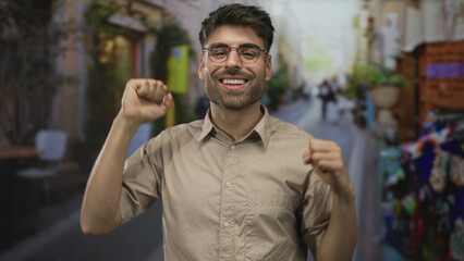 Obraz premium Smiling bearded man wearing glasses clenches fists on a sunny city street with shops and passerby blur; joy triumph.