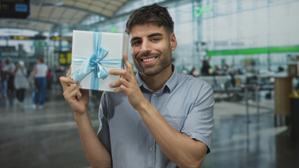 Obraz premium Man holds wrapped gift box near face in busy airport terminal while smiling at camera; joy anticipation celebration.